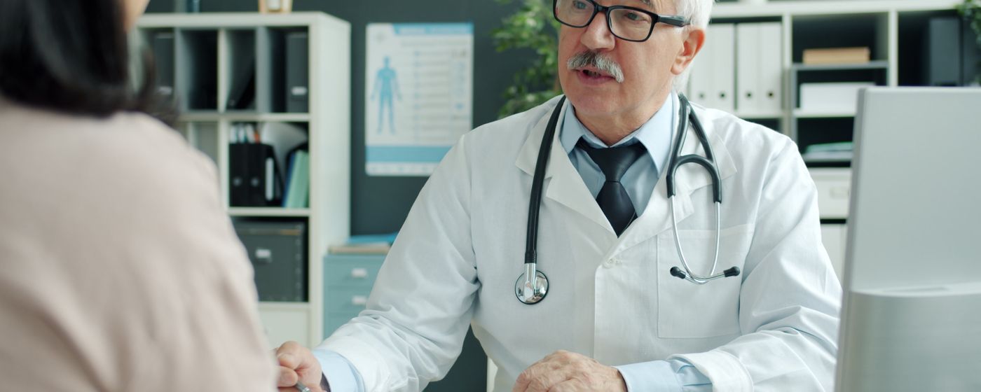 A doctor in a white coat and stethoscope talking with a patient across a desk in a clinic