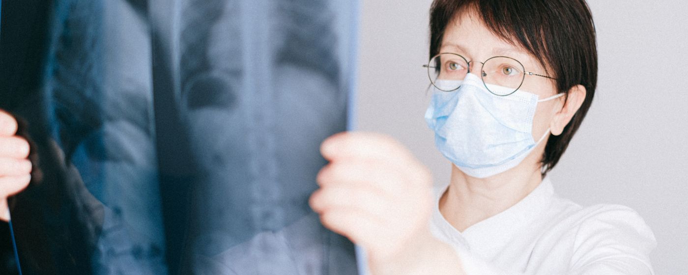 A clinician in a face mask holding an X ray film up to the light to read it