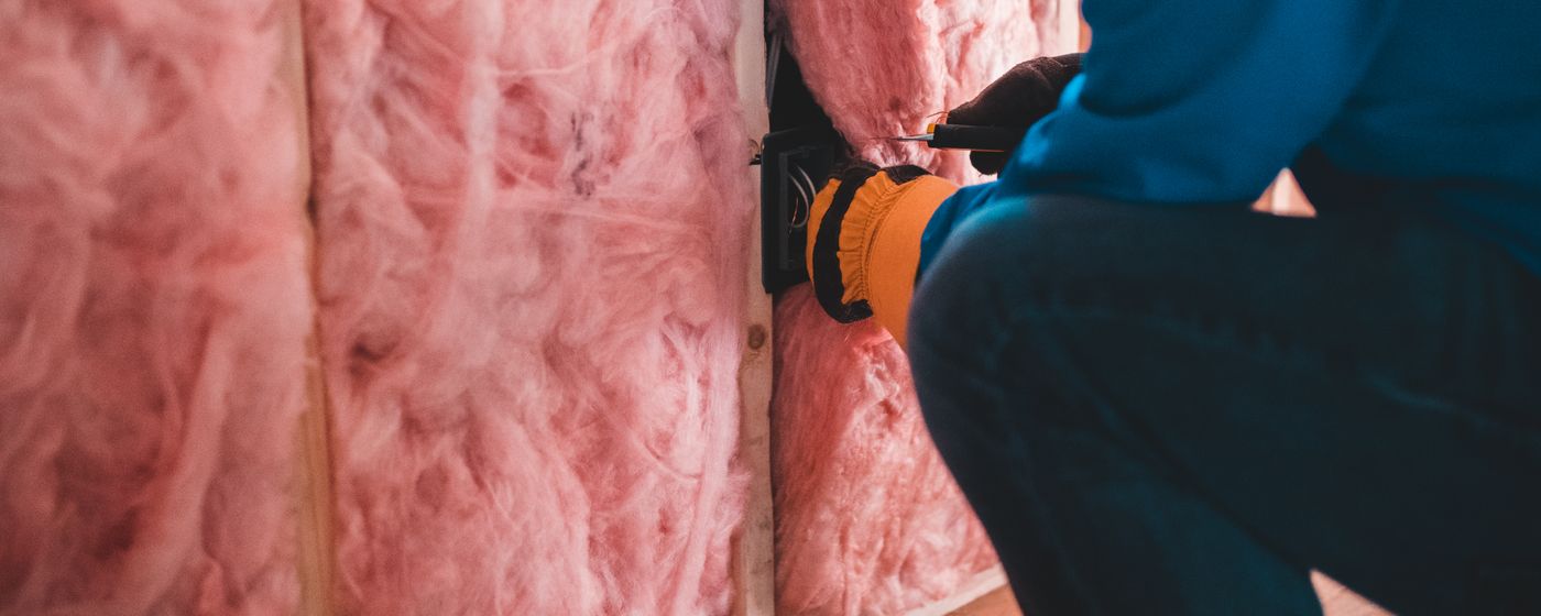 A person kneeling beside pink fiberglass insulation in an unfinished wall