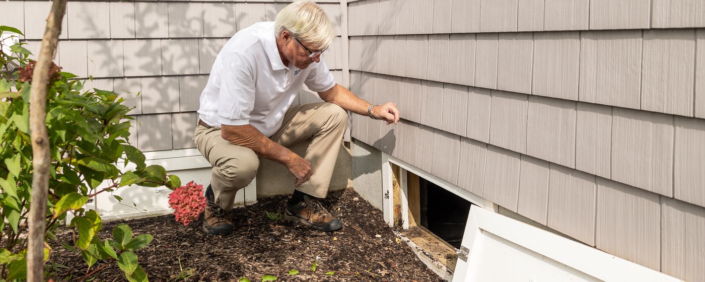 An older man crouching beside the foundation vent of a shingled house