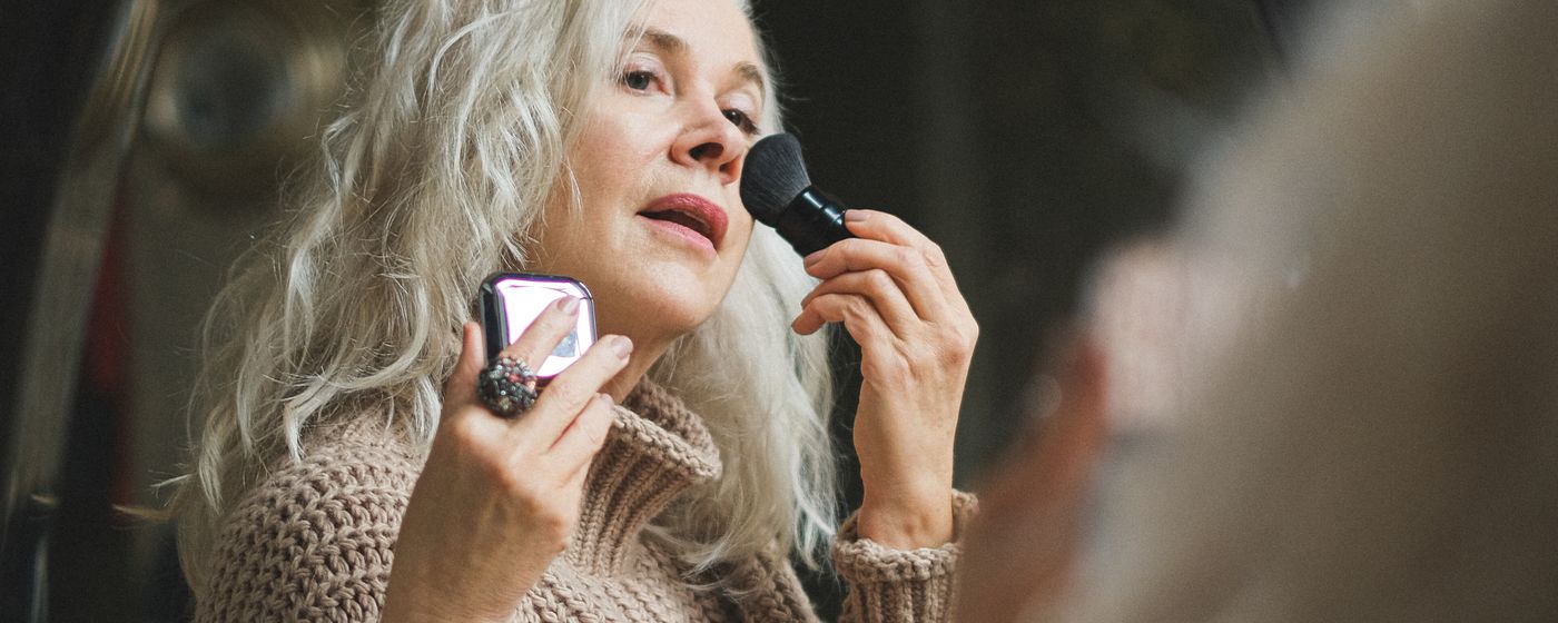 An older woman applying makeup to her cheek in front of a bathroom mirror