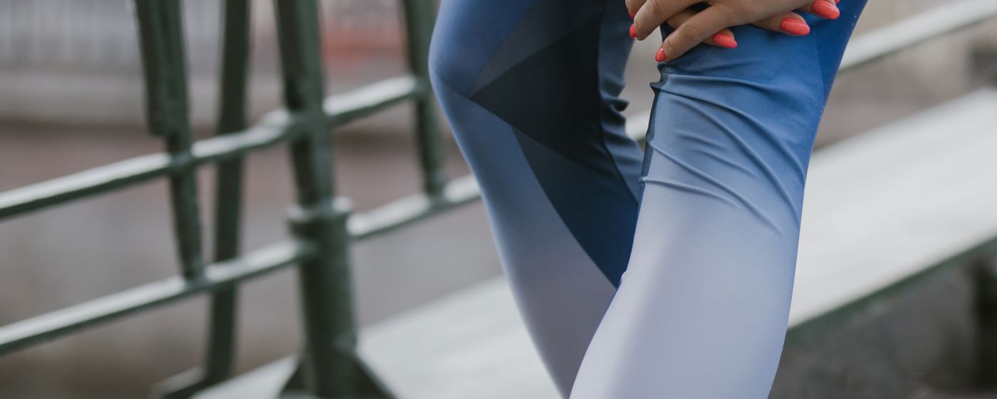 A person in blue leggings stretching a raised leg against a metal rail outdoors