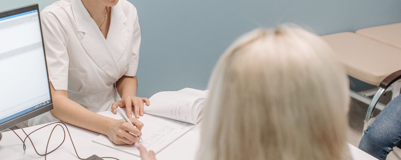 A clinician writing notes at a desk while sitting with a patient in a consultation room