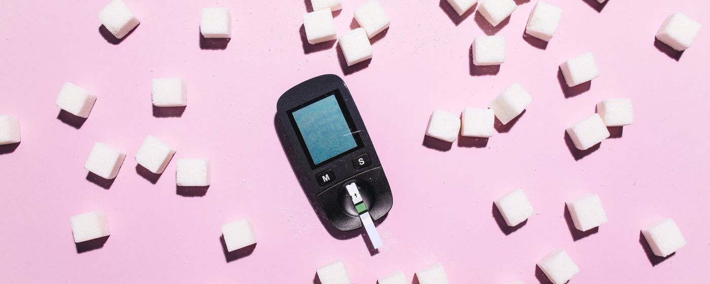 A blood glucose meter lying on a pink surface surrounded by scattered sugar cubes