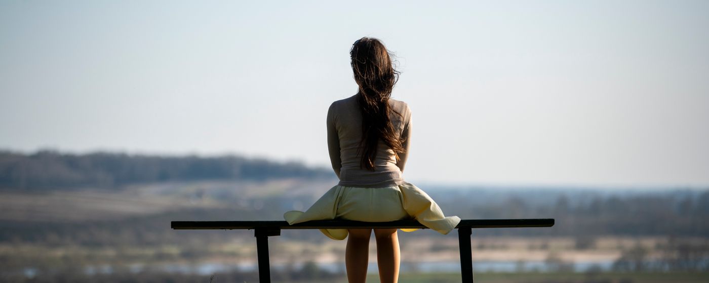 A person sitting alone on a bench facing an empty winter field under a pale sky