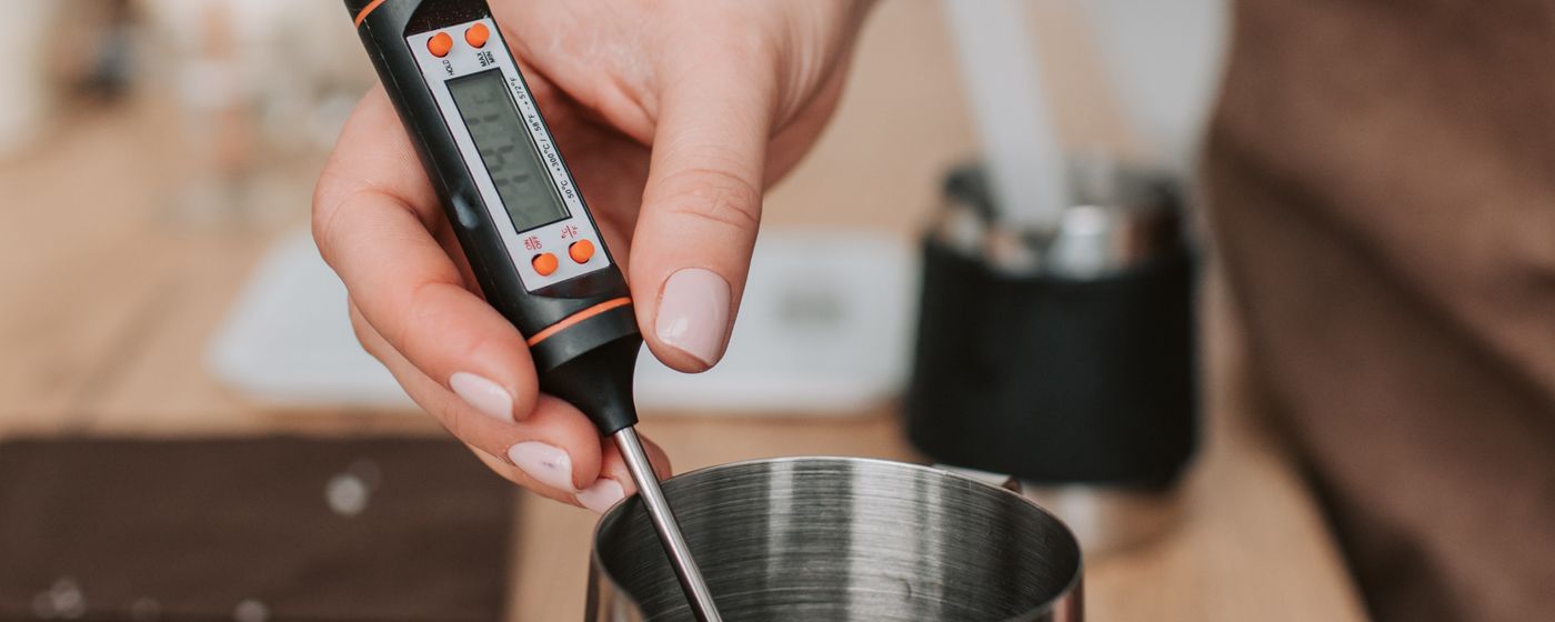 A hand holding a digital probe thermometer in a small metal pot on a kitchen counter