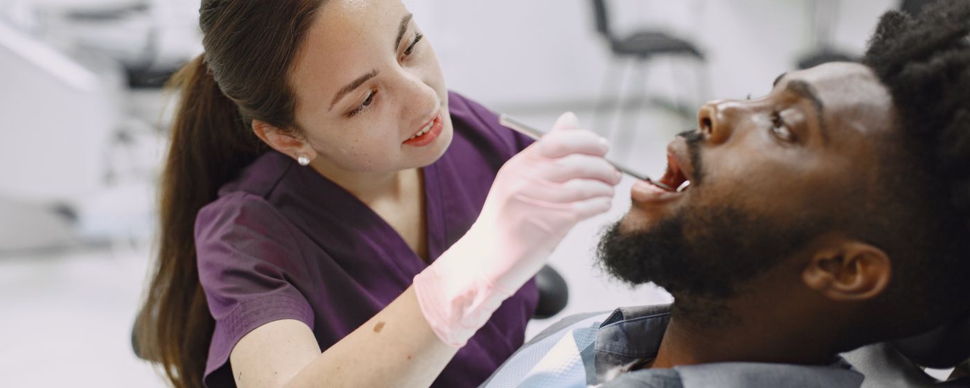 A dental hygienist in purple scrubs examining a seated man's open mouth in a clinic