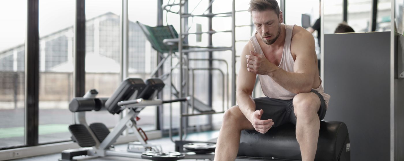 A man in a tank top sitting on a gym bench holding his shoulder after a workout