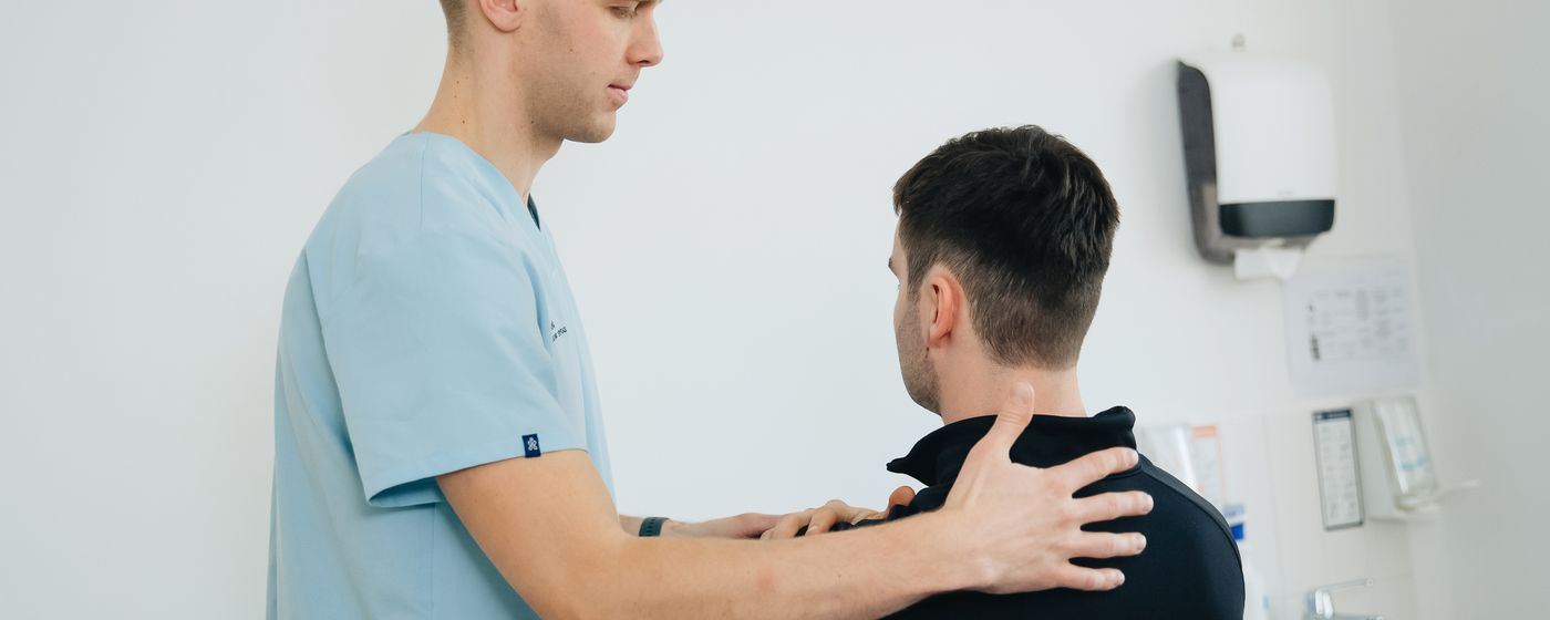 A physiotherapist in scrubs assessing the shoulders of a seated patient in a clinic