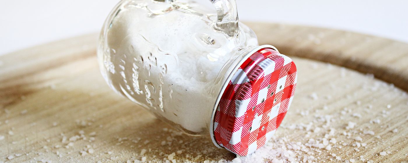A glass jar tipped over on a wooden table with salt spilling out of it