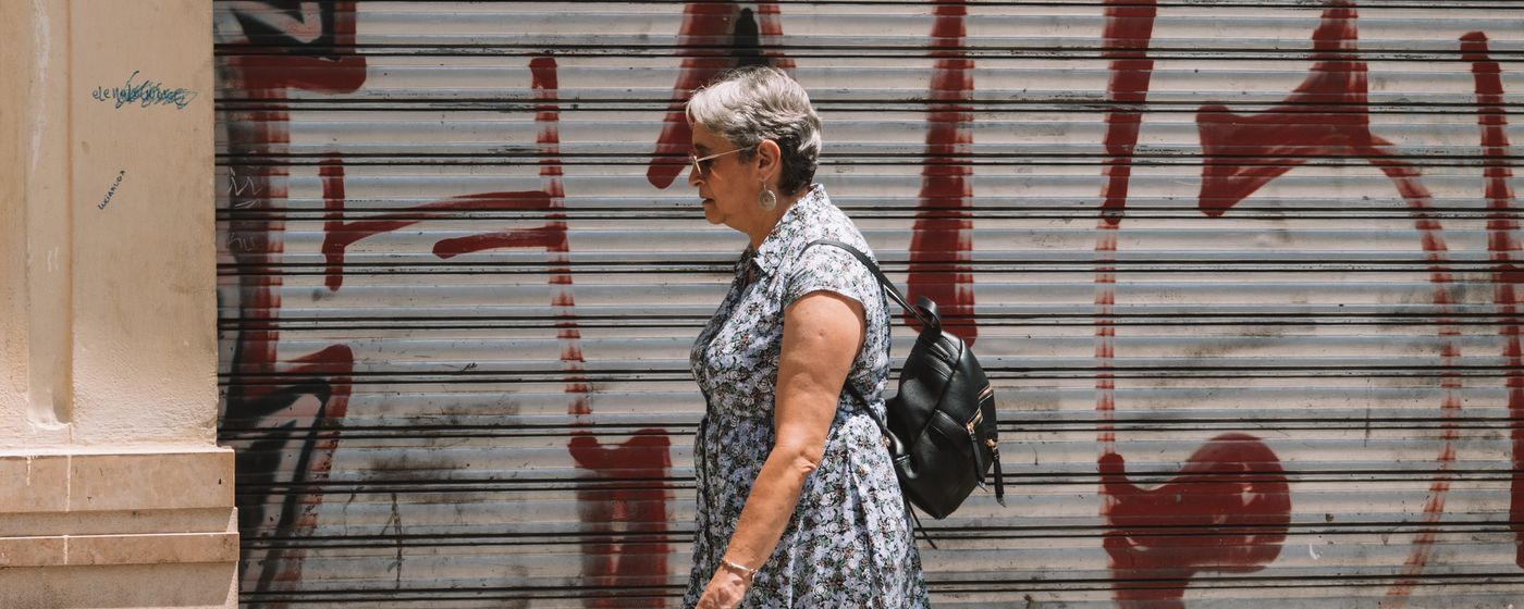 Older woman walking along a city street past a metal shutter, carrying a small backpack