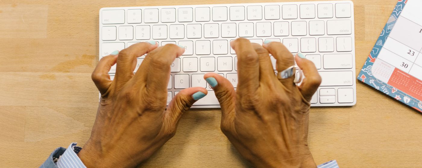 Older adult hands resting on a white keyboard on a wooden desk, seen from above