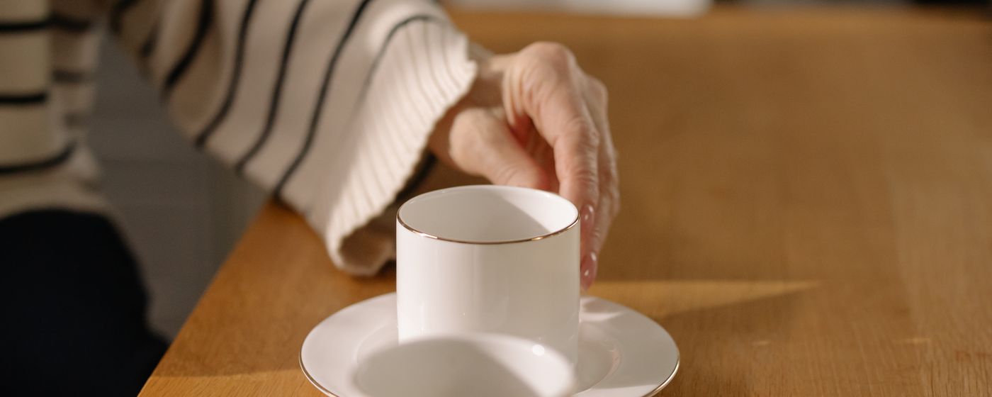 Older adult reaching for a white coffee cup and saucer on a wooden table