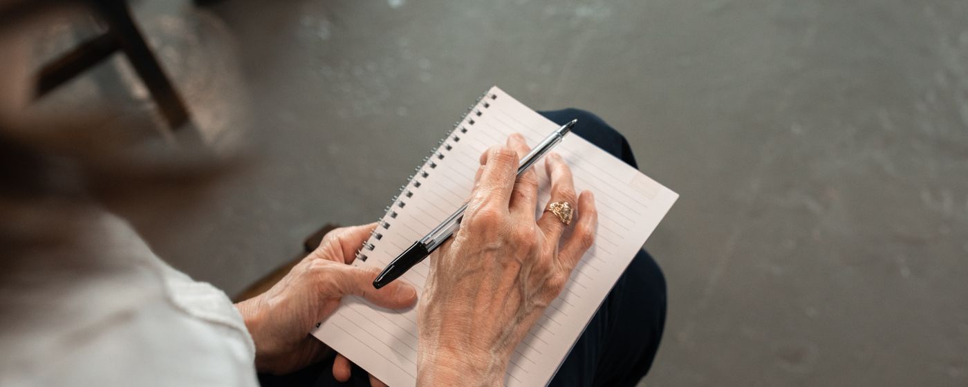 Older adult writing in a spiral notebook with a pen, hands and paper filling the frame