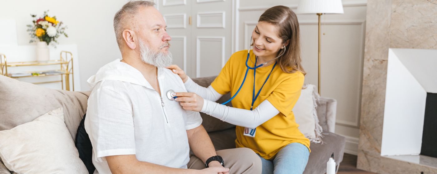 A nurse uses a stethoscope during a home check-up with a senior man on a couch