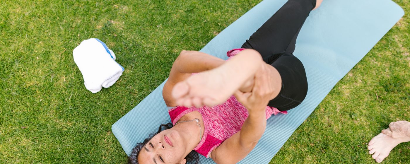 A woman on a yoga mat in a park pulling one knee toward her chest