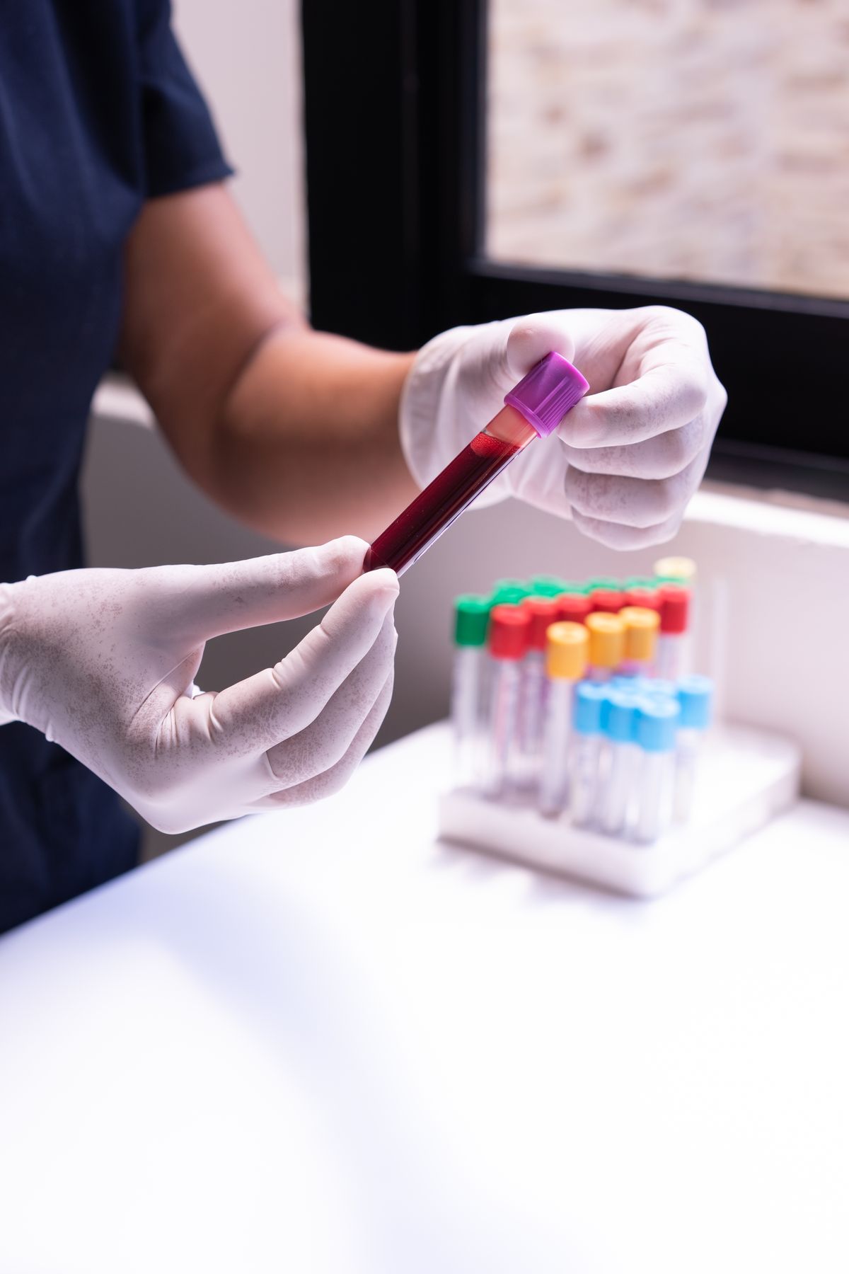 Gloved hands holding a filled blood collection tube beside a rack of coloured sample tubes