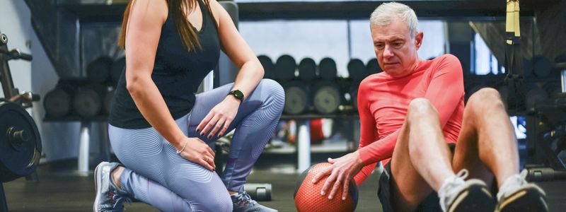 A senior man working with a personal trainer on a strength exercise in a gym
