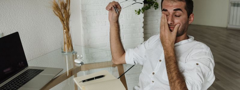 A man yawning at a desk beside an open laptop during the day