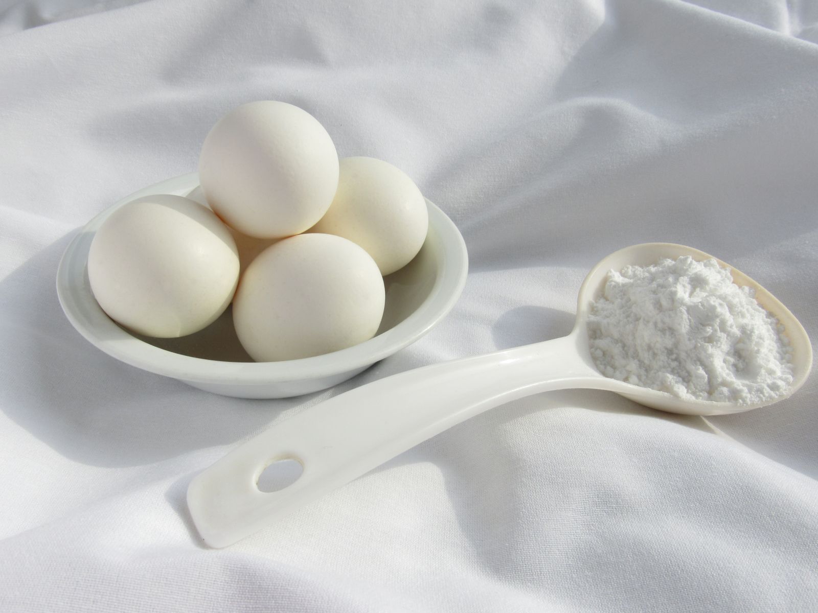 A bowl of whole eggs beside a spoon of white powder on a white cloth