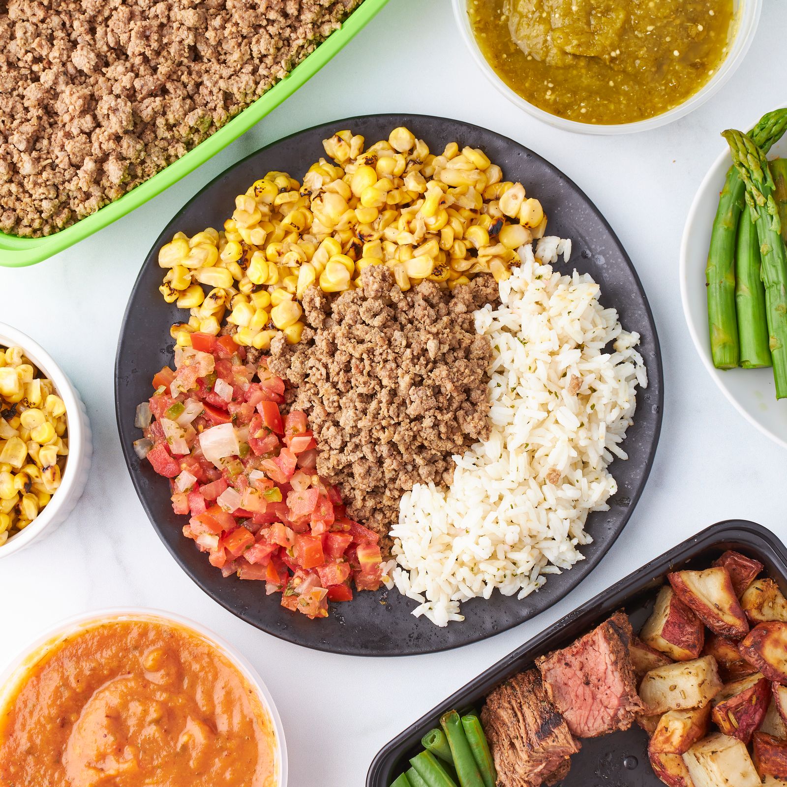 Cooked ground beef, rice, corn and salsa portioned on a dark plate beside prepared containers