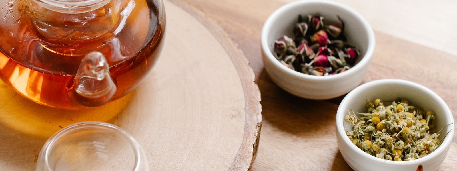 Small ceramic bowls of dried herbs and flowers beside a glass teapot on a wooden board