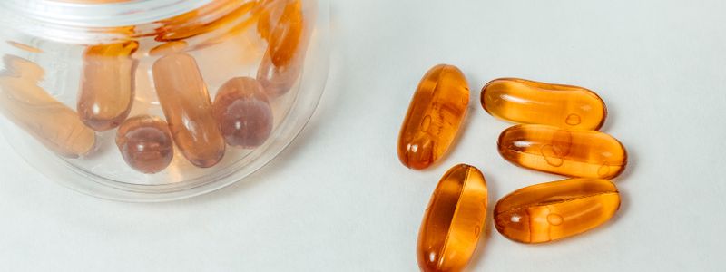 Amber supplement capsules spilling from a clear jar on a pale surface