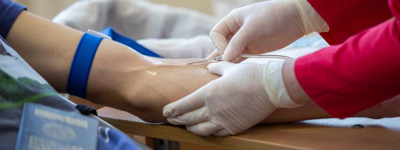 A gloved clinician drawing a blood sample from a person's arm with a blue tourniquet