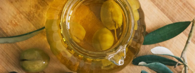 An overhead view of a glass cruet of golden olive oil beside fresh olives and olive leaves on a wooden board