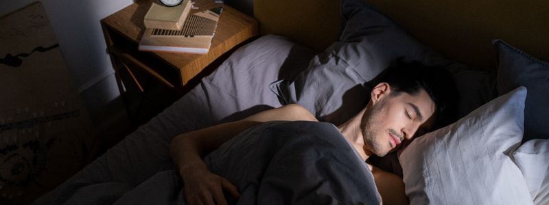 A man asleep on his side in a dimly lit bedroom beside a wooden bedside table