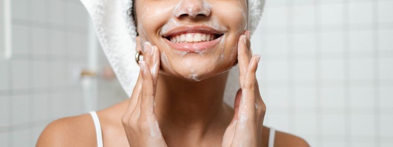A smiling woman with a towel wrapped around her hair lathering a white cleanser across her cheeks