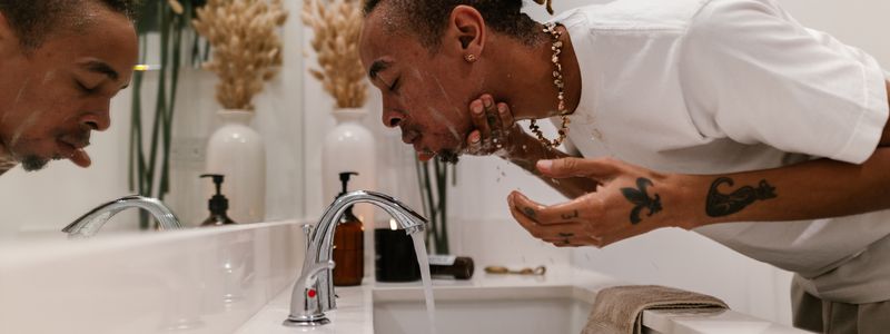 A man leaning over a white bathroom sink rinsing his face with running water