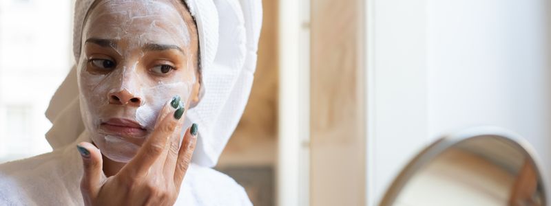 A woman with a towel on her hair spreading white moisturizing cream across her cheek