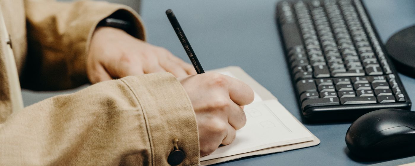 Hands writing in a small notebook on a desk beside a keyboard