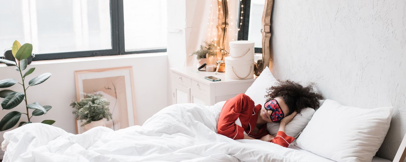 Woman lying in a white bed wearing a sleep mask in a bright bedroom with plants