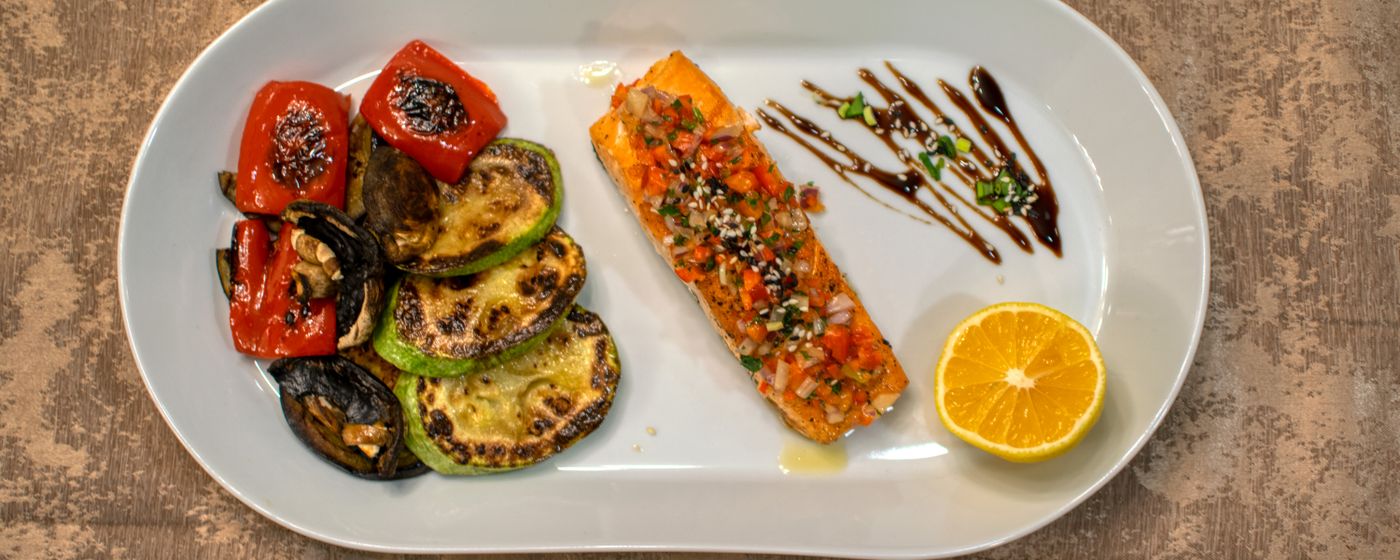 White plate of grilled salmon with roasted peppers, courgette and a lemon half