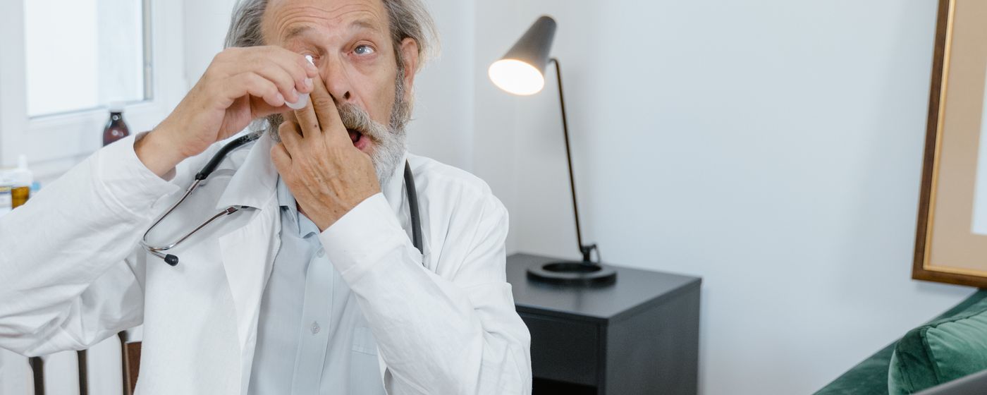 A senior doctor sits at a desk applying eye drops