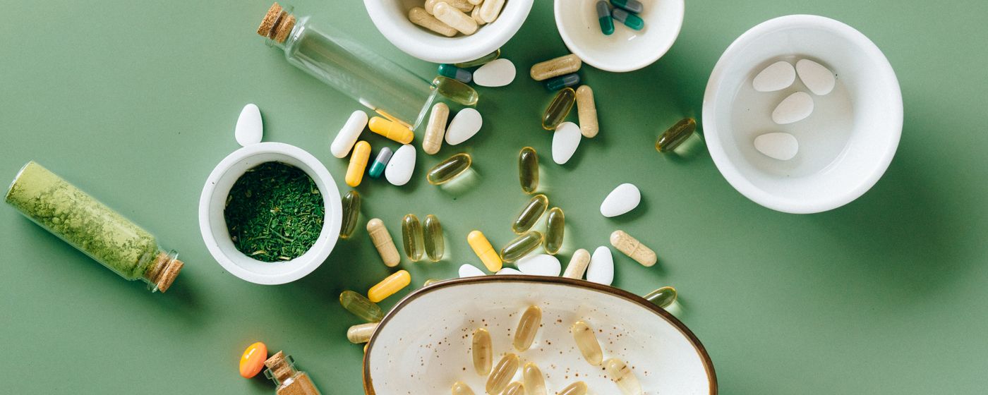 Overhead view of assorted vitamin capsules and tablets in small bowls on a green background