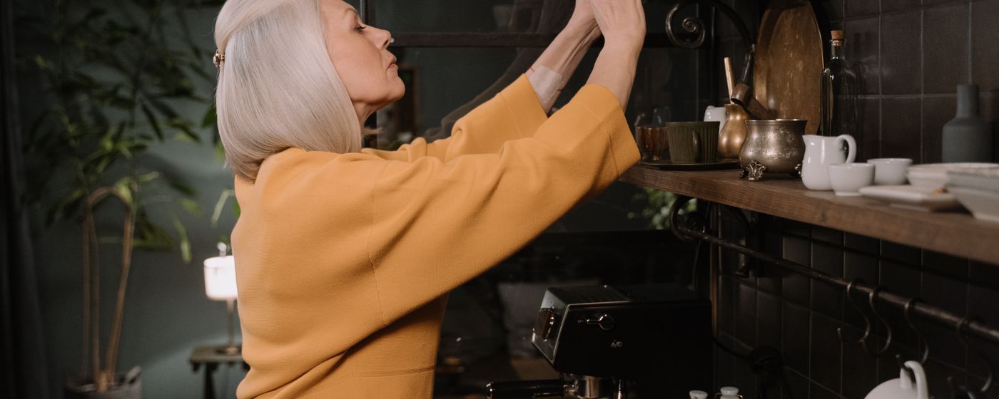 An older woman with white hair reaching up to a high kitchen shelf