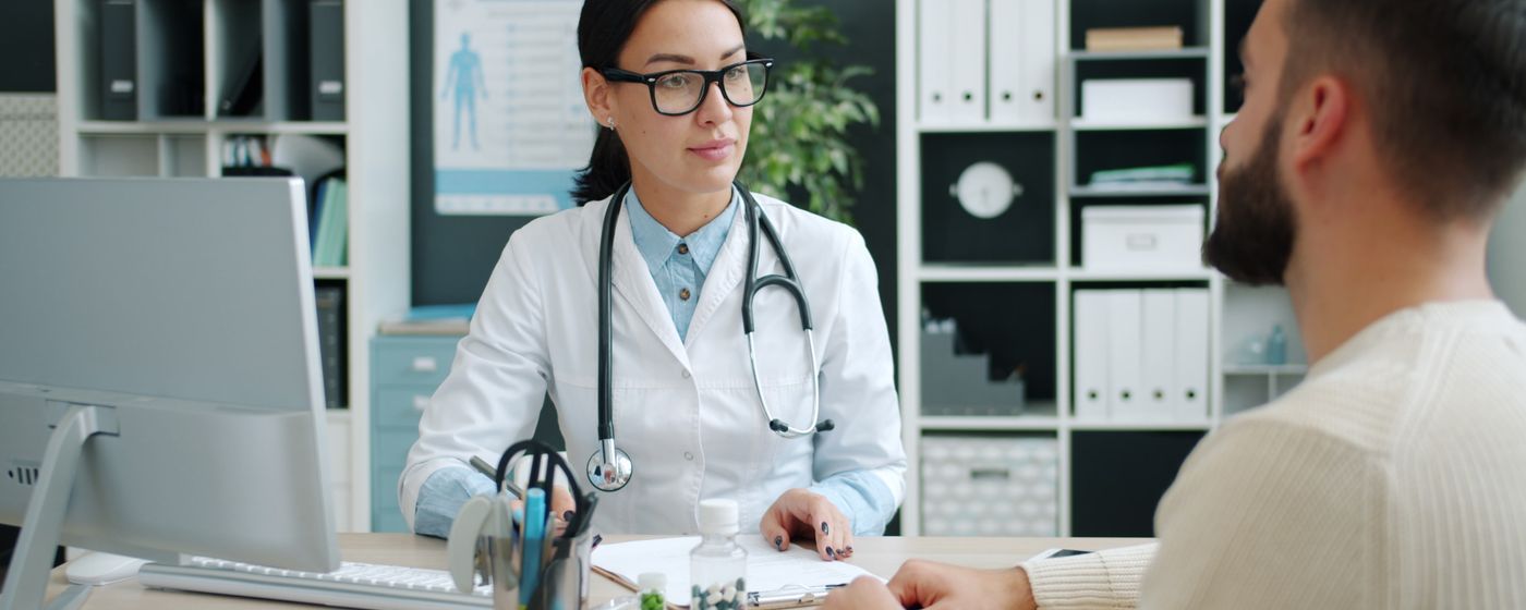 A clinician in a white coat with a stethoscope talking with a patient across a desk