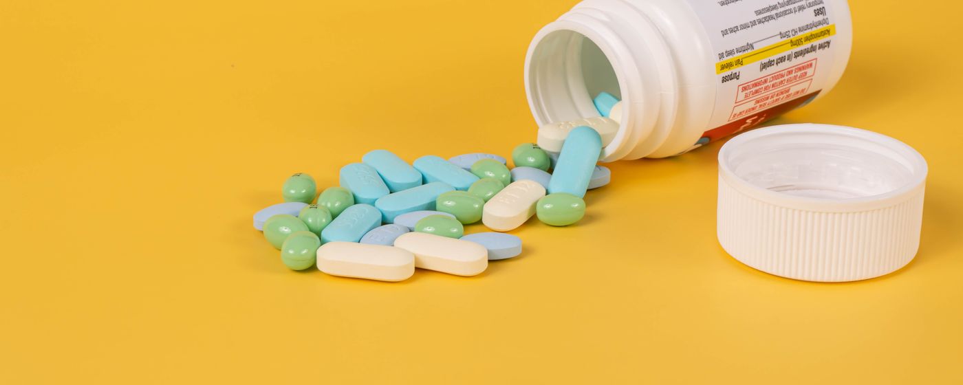 A tipped over pill bottle with coloured tablets and capsules spilled on a yellow surface