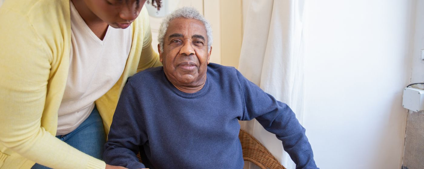 A seated older man with a carer's hand resting on his shoulder