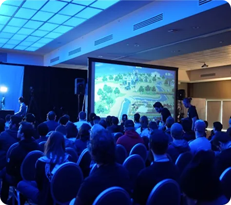 Audience watching a video game displayed on a large screen in a dimly lit room.