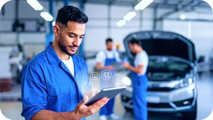 Mechanic in blue jumpsuit using a tablet with two other mechanics and a car with open hood in the background inside a garage.