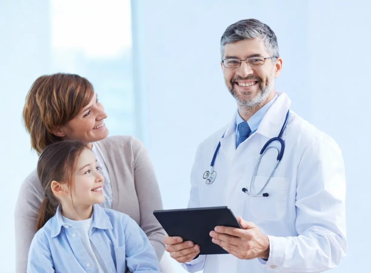 Smiling male doctor holding a tablet with a mother and her young daughter in a bright medical office.