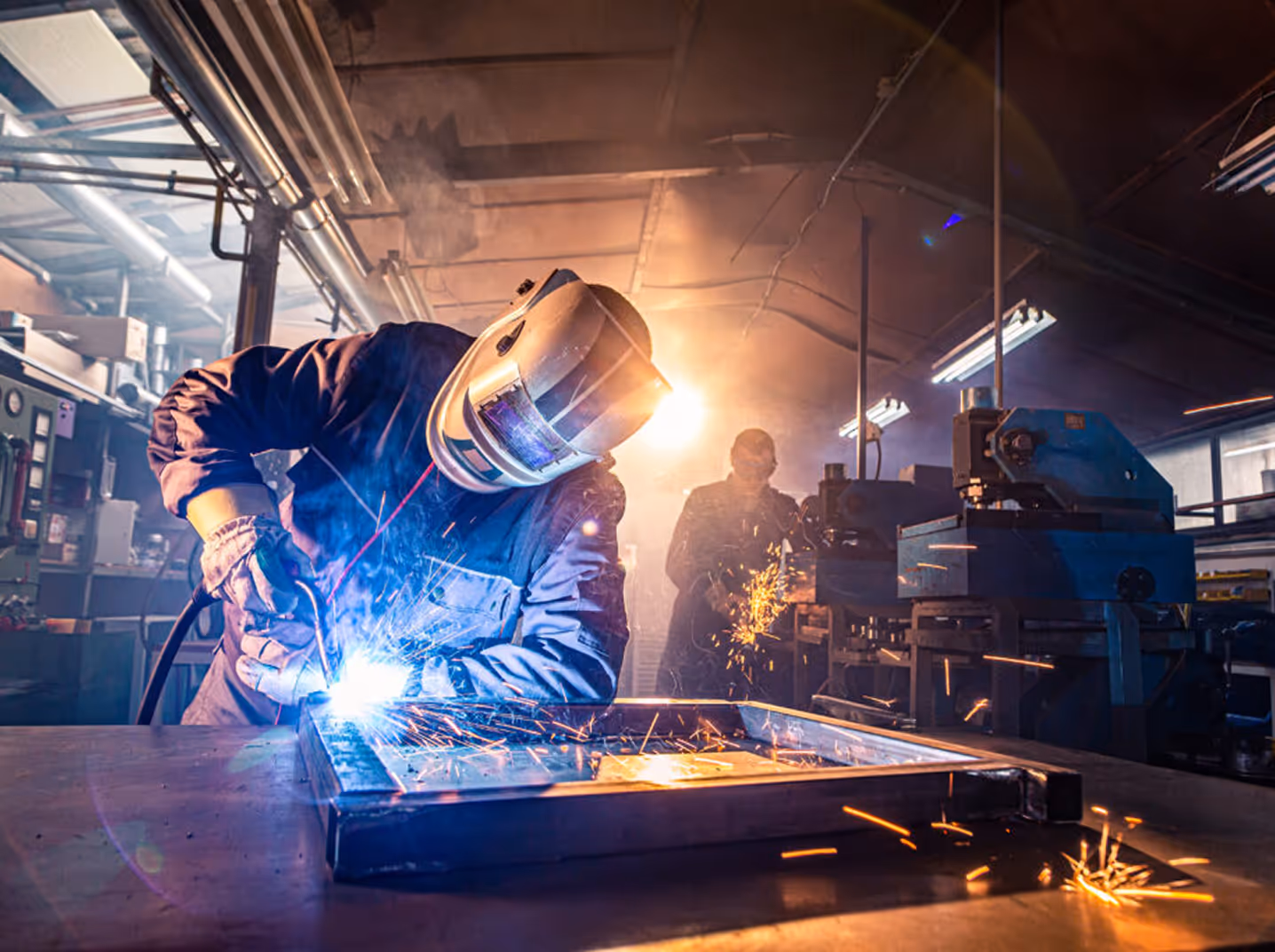 Worker welding metal frame with protective helmet in an industrial workshop with sparks flying.