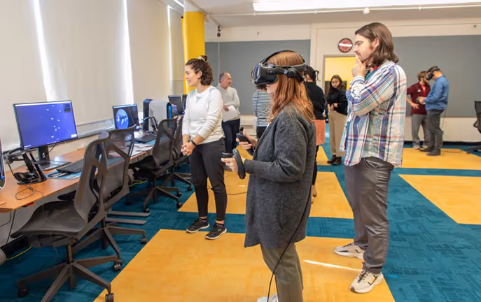Person using virtual reality headset with controllers in a room with multiple people and computer workstations.
