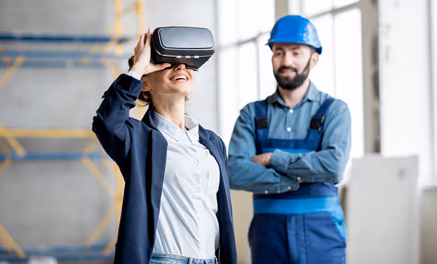 Woman wearing VR headset standing next to a man in a blue hard hat and work overalls inside a construction site.