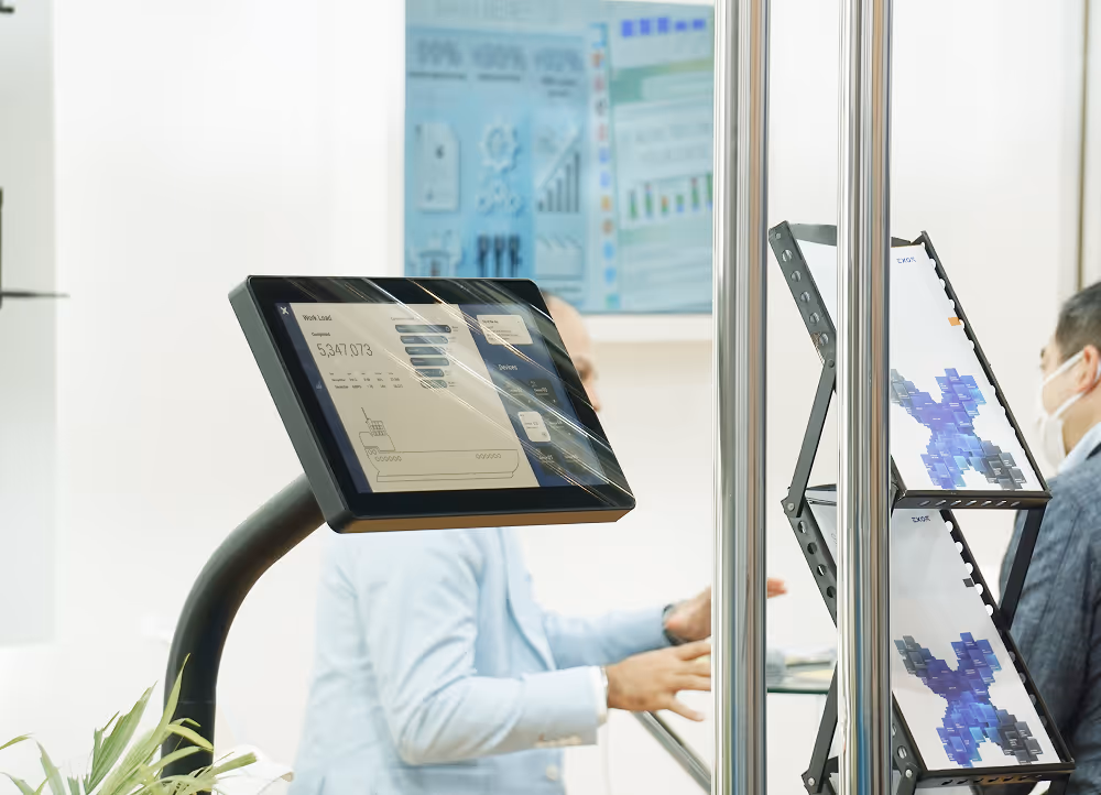 Touchscreen monitor displaying data charts in front of two men in suits having a discussion, with brochures on a stand nearby.