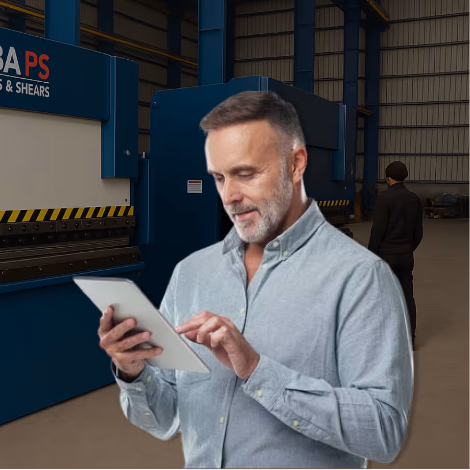 Man with grey beard in light shirt using a tablet in a factory with industrial machinery in the background.